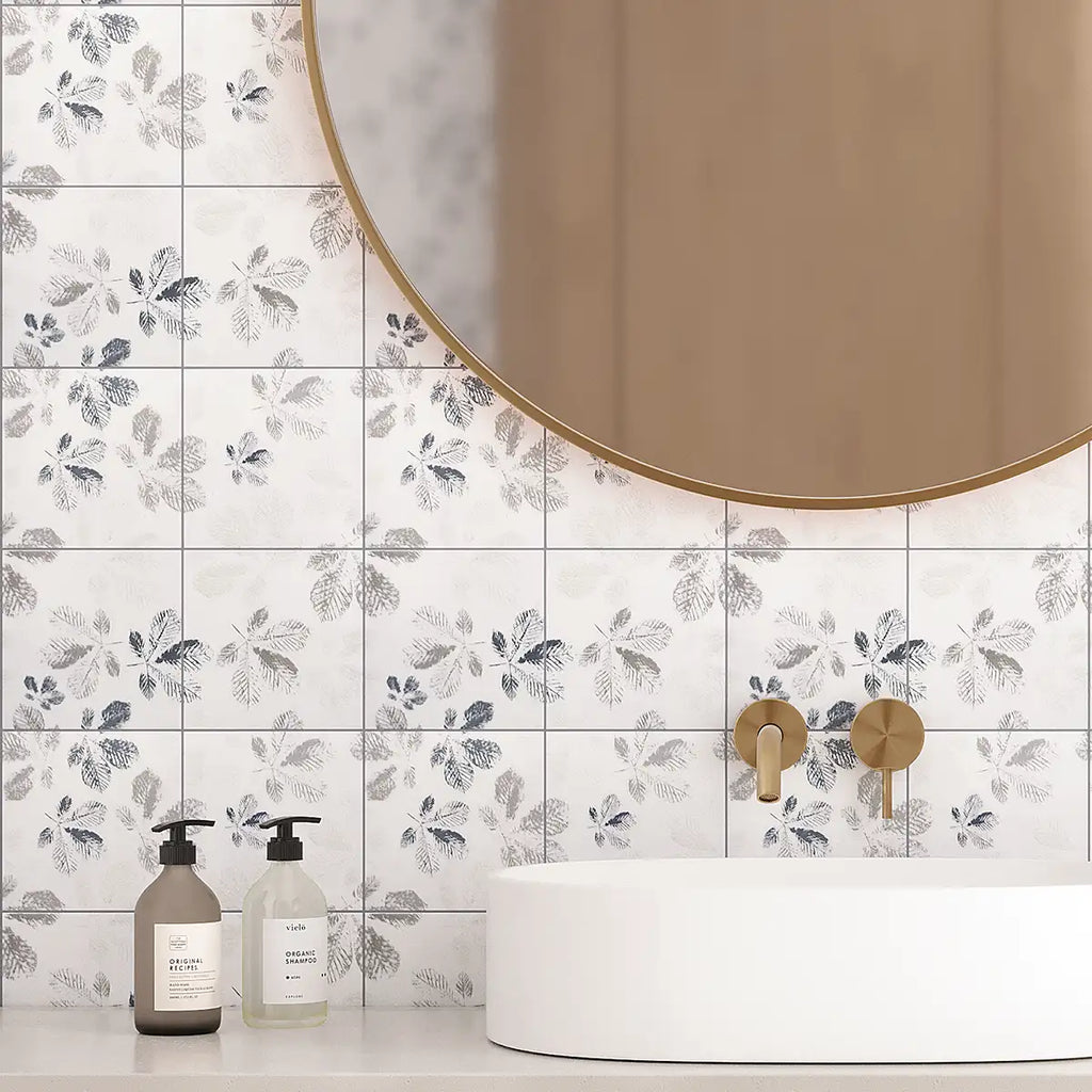 White square wall tiles with a gray botanical leaf pattern, installed in a bathroom setting behind a white vessel sink and a round gold-framed mirror.