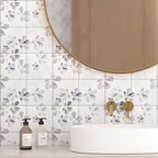 White square wall tiles with a gray botanical leaf pattern, installed in a bathroom setting behind a white vessel sink and a round gold-framed mirror.