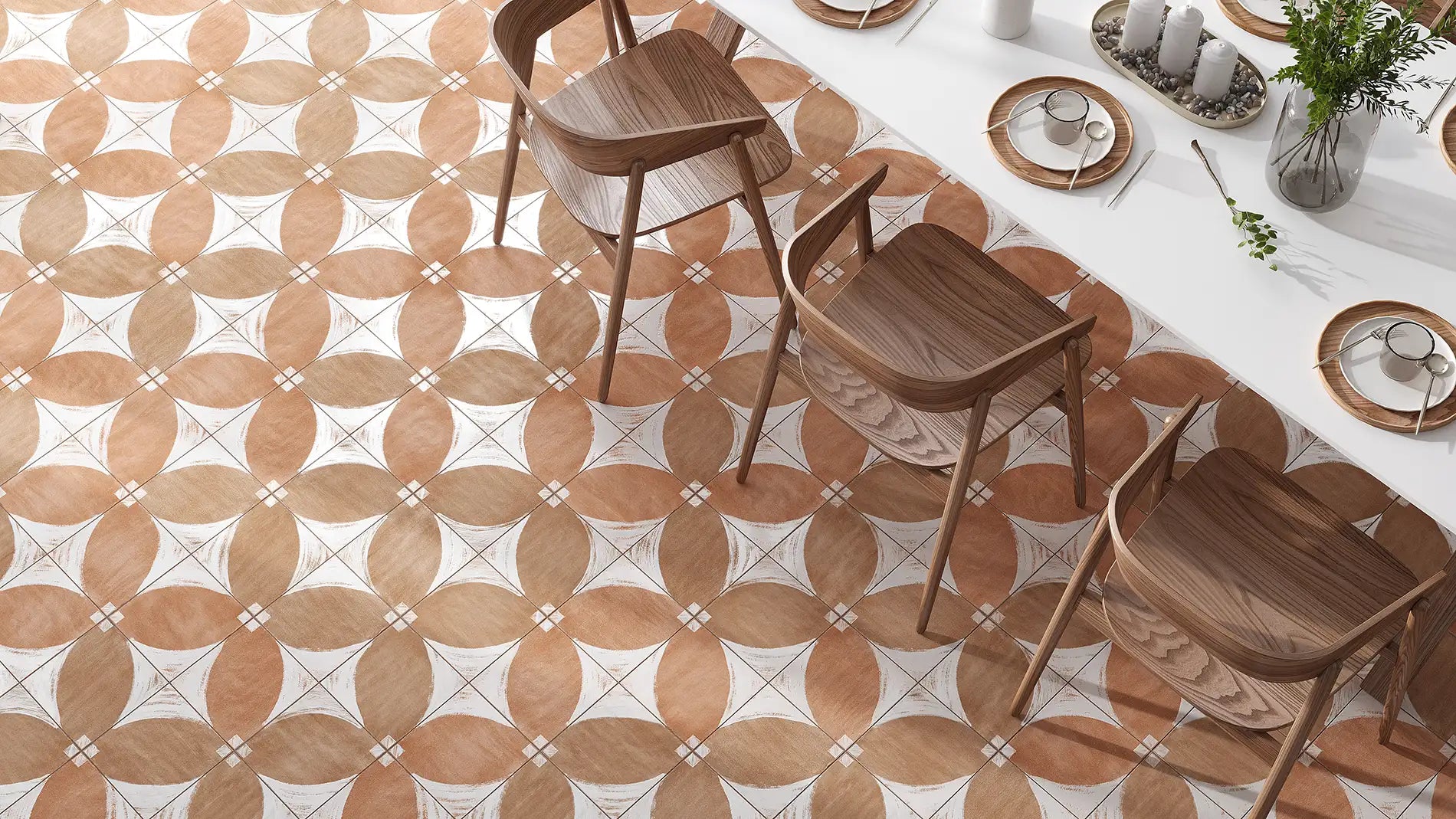A top-down view of a dining room floor featuring a geometric patterned tile design in warm terracotta, beige, and white. Three wooden dining chairs are arranged on the floor next to a white table set with plates and candles.